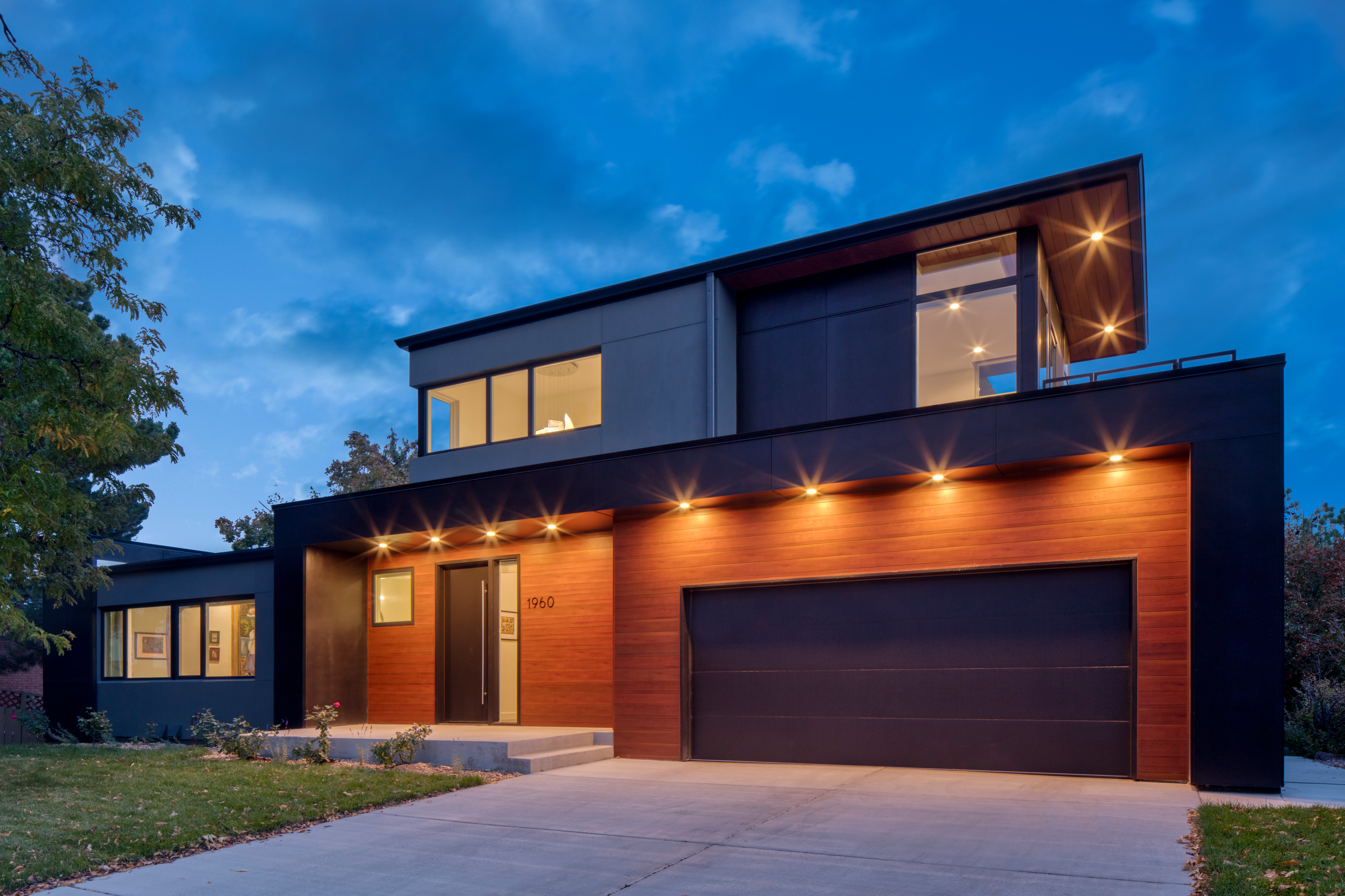 Evening view of a modern two-story Petra Custom Builders home with illuminated exterior sconces and a warm wooden garage door