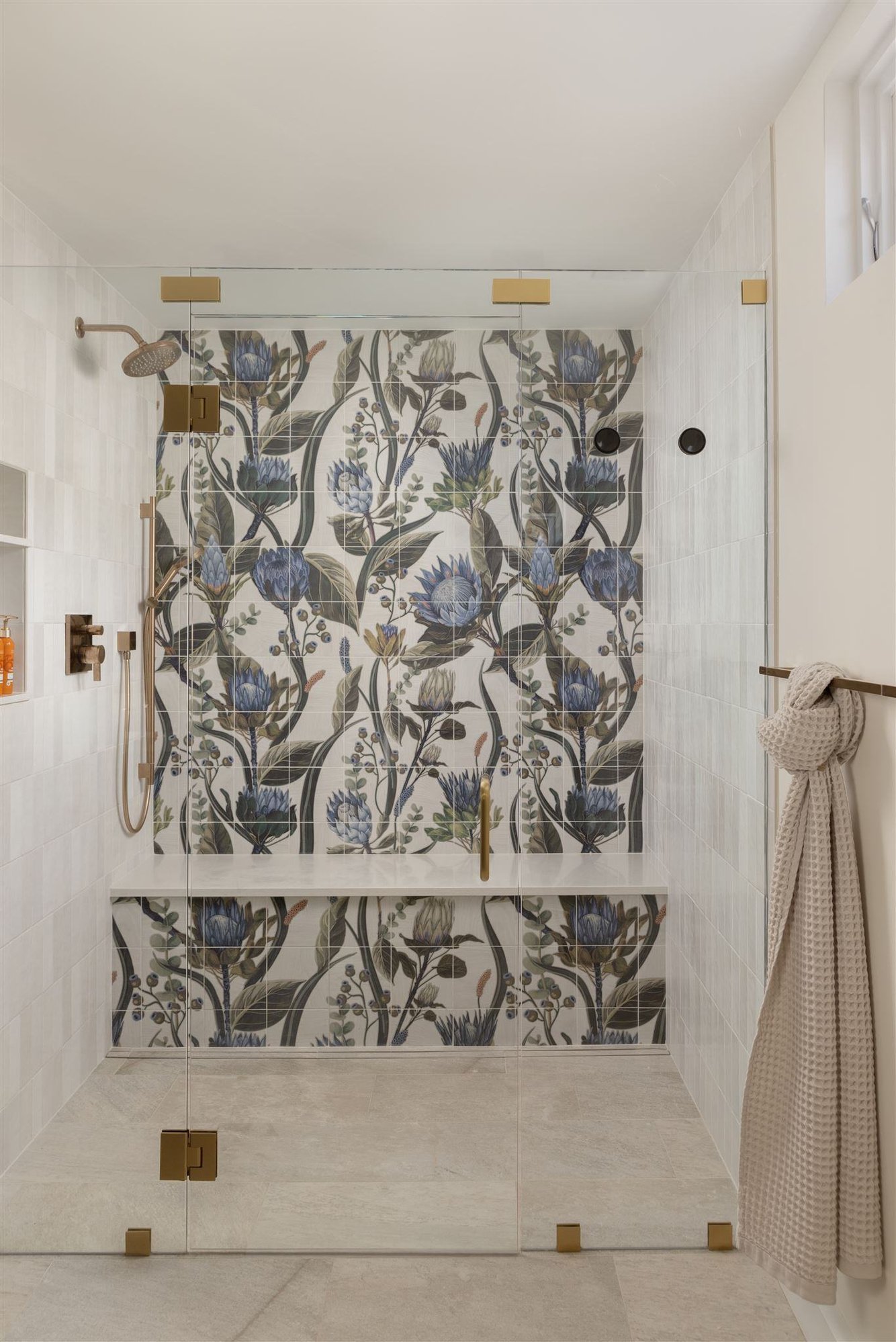 Shower with floral mural style tile, frameless glass shower door, and gold hardware