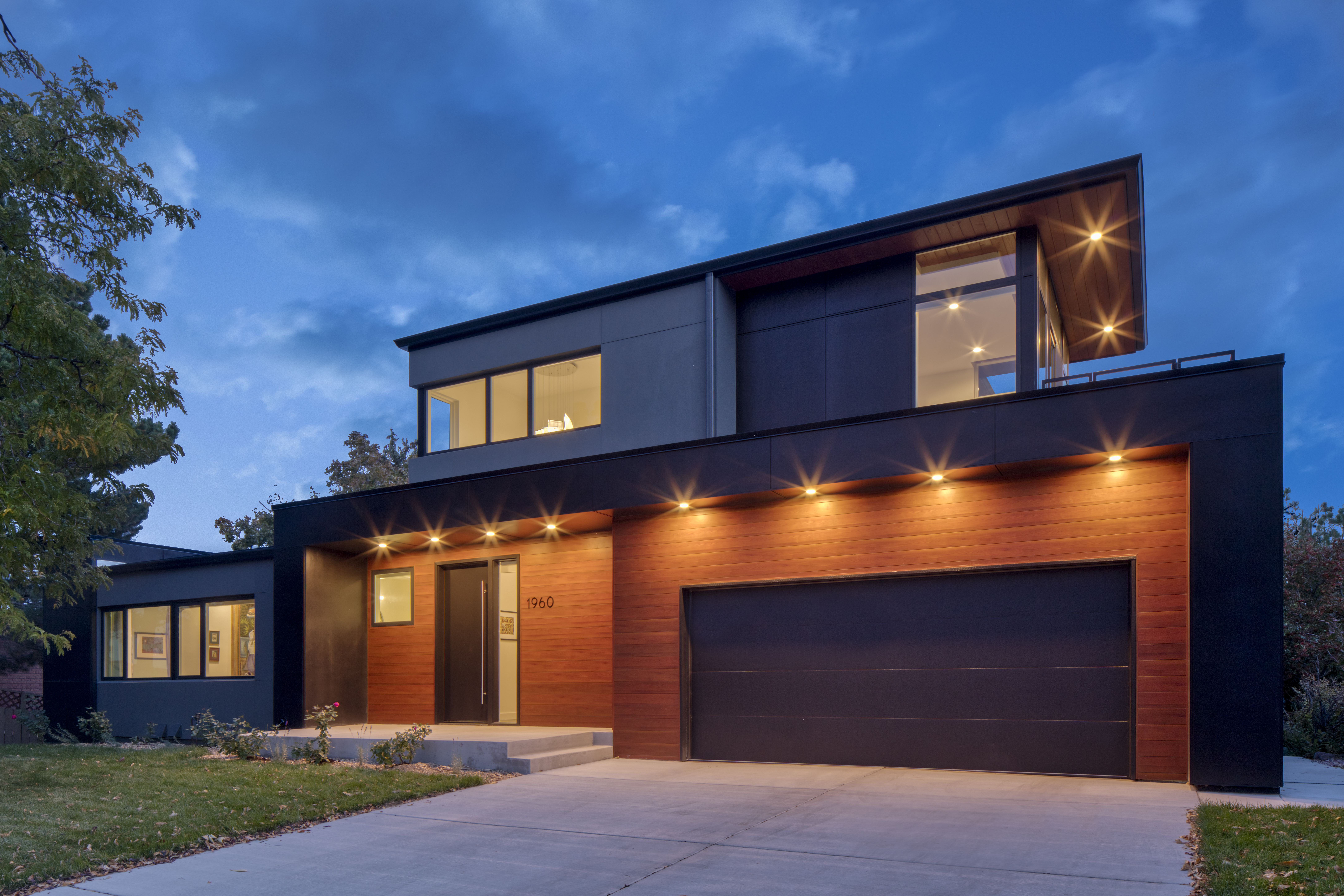 Evening view of a modern two-story Petra Custom Builders home with illuminated exterior sconces and a warm wooden garage door